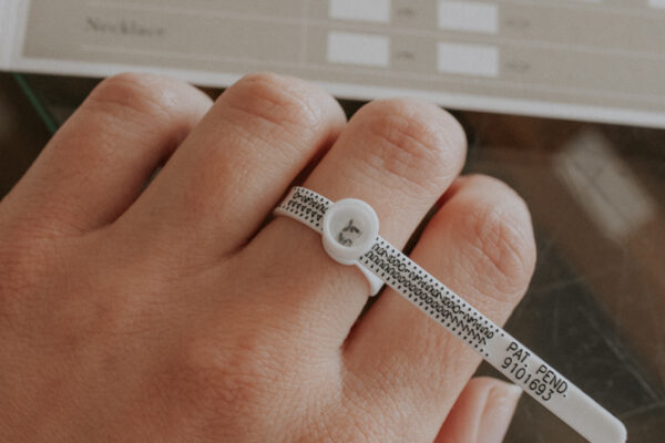 een hand waarop aan één vinger de ringsizer wordt gebruikt om te demonstreren hoe je je ringmaat op kan meten, en de ringsize guide op de achtergrond